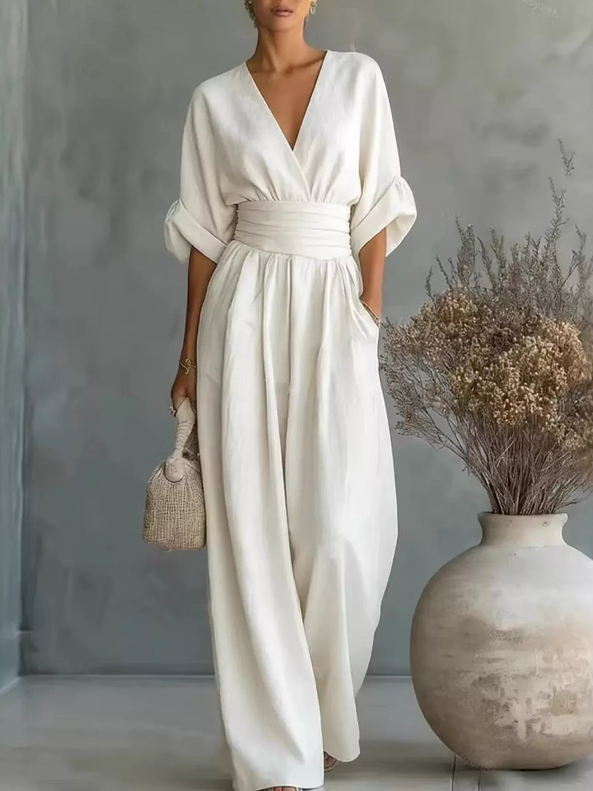 Woman in a white dress standing next to a vase with dried flowers against a gray wall.
