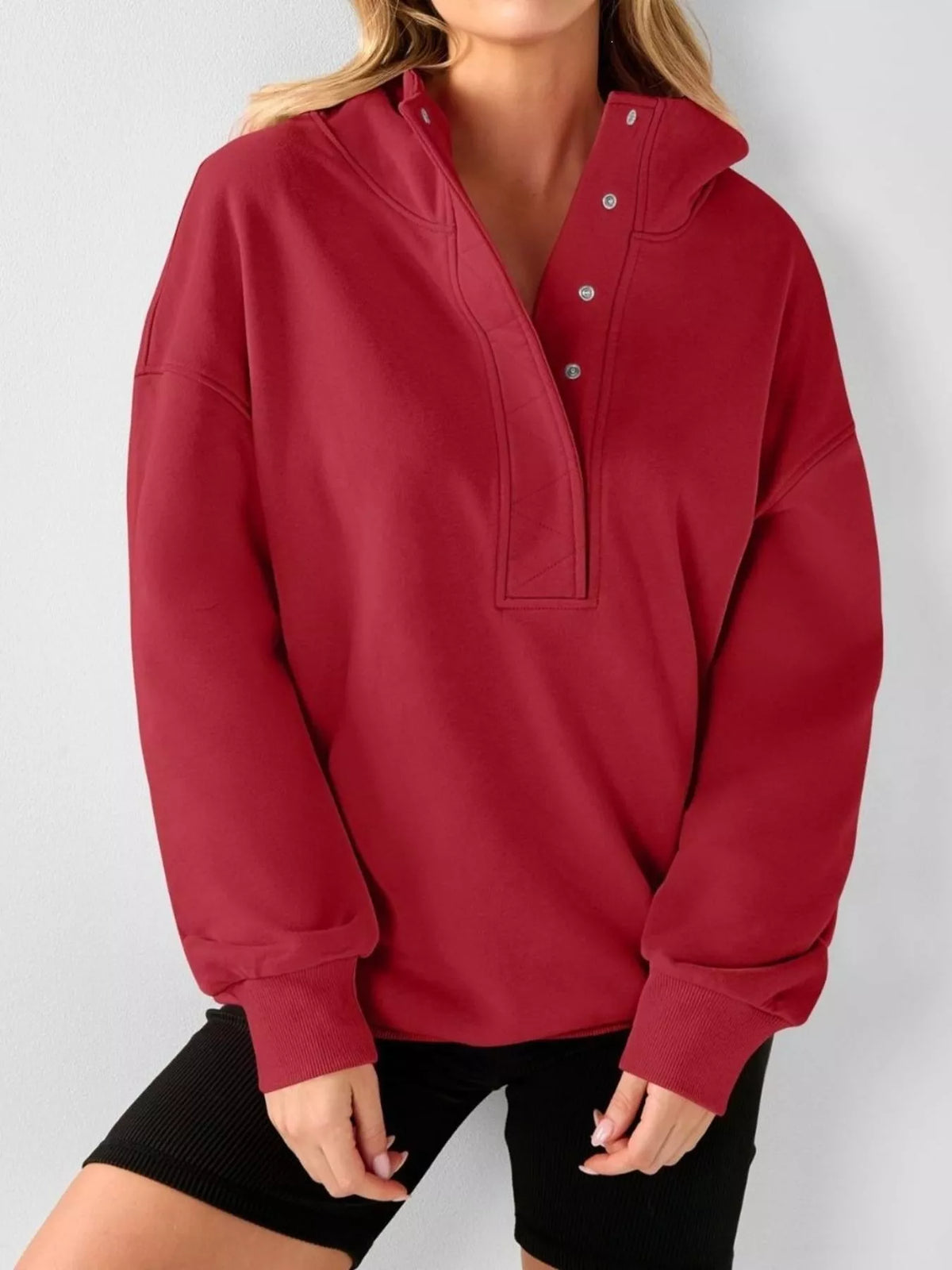 A woman wearing a red long-sleeve half-button pullover top with a mock neck and black shorts, shown from the neck down against a light gray background.