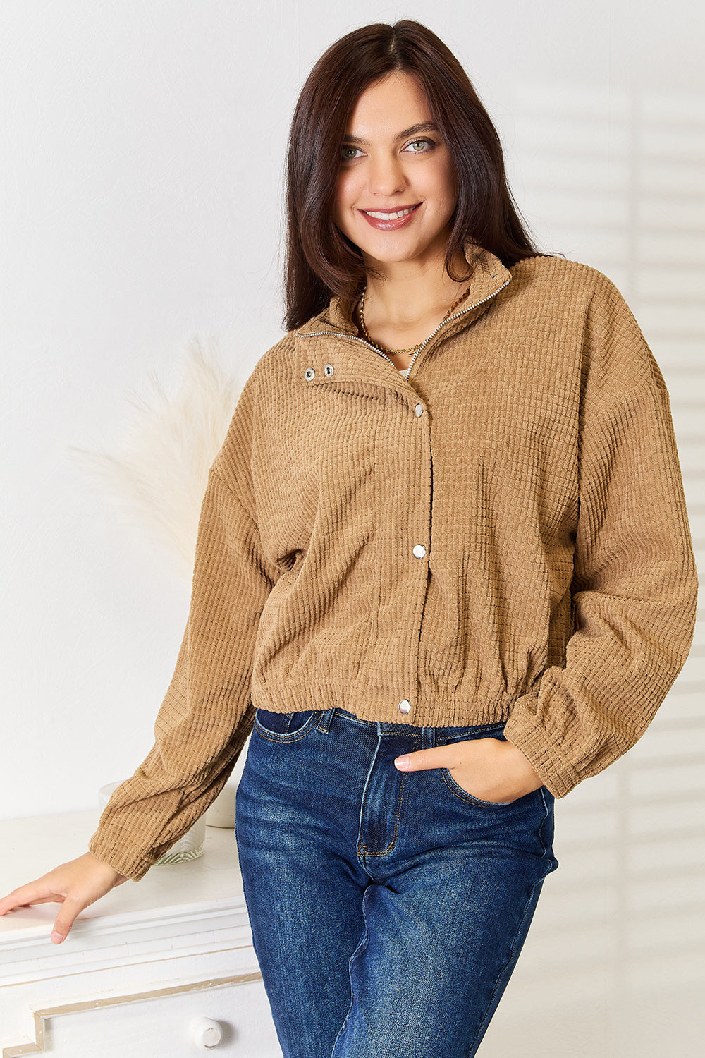 A woman wearing a tan waffle-knit zip-up jacket with a mock neck and silver snap buttons, paired with dark blue jeans, posing indoors against a light-colored wall.