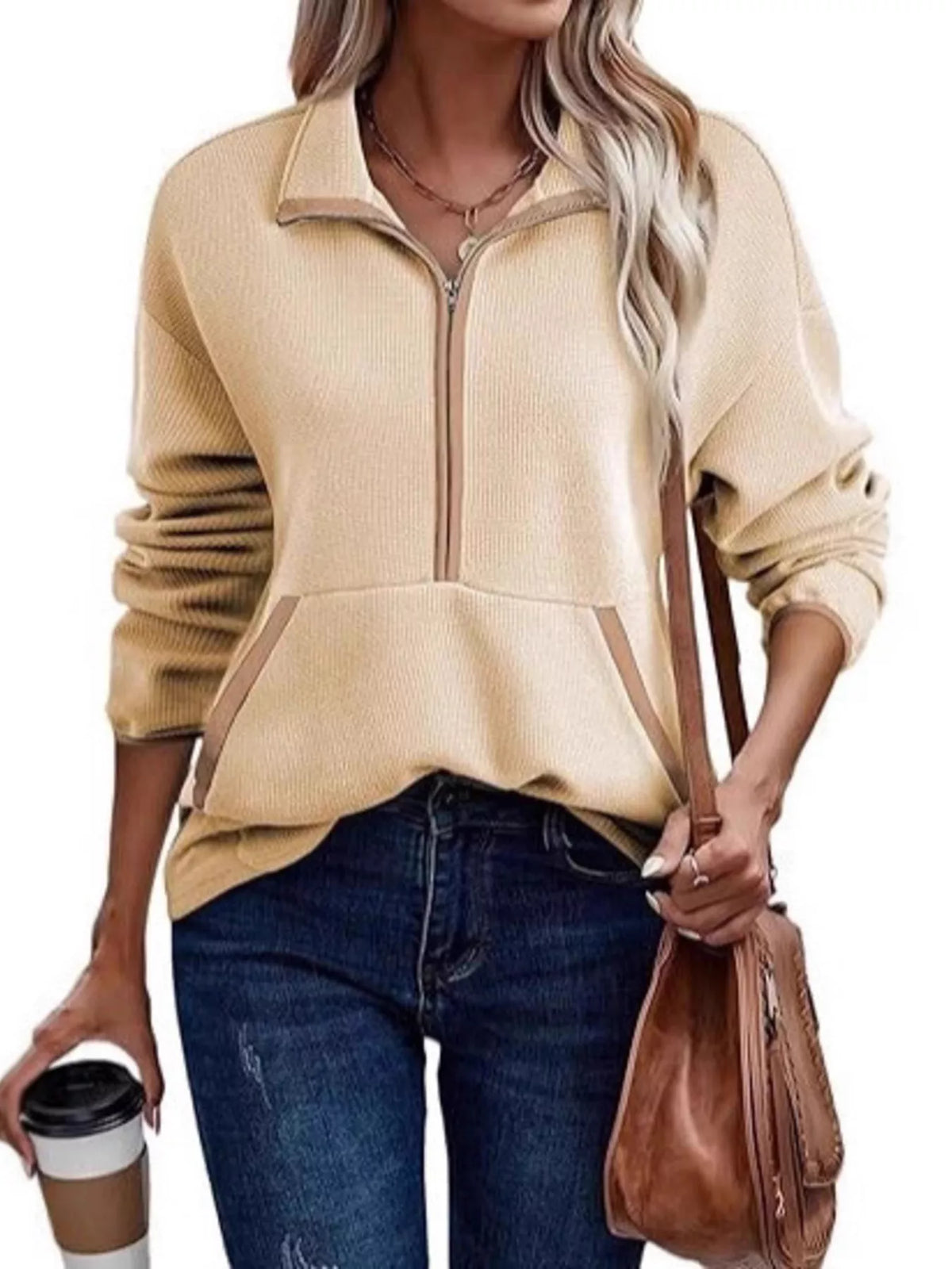 Woman wearing a beige zip-up sweater with blue jeans, holding a brown bag and a coffee cup.