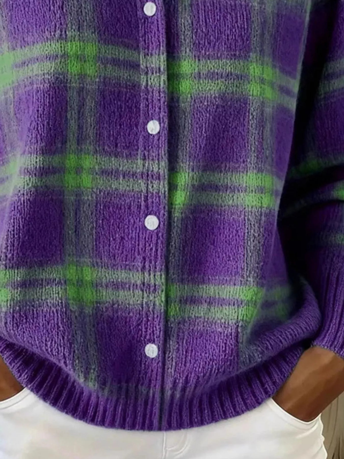 A close-up of a person wearing a fuzzy knit cardigan with a purple and green plaid pattern, white buttons, and white pants.