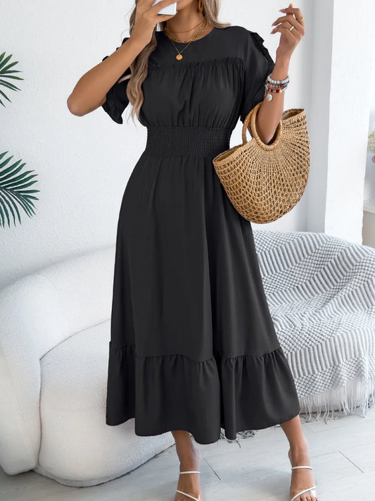 Woman wearing a black dress holding a straw bag in a room with a white couch and decorative plant.