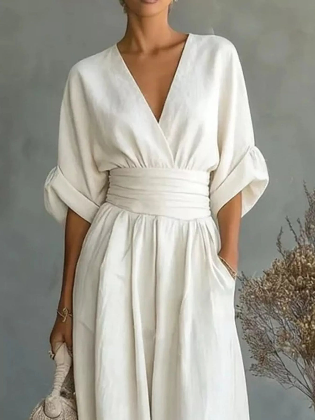 A woman wearing a cream-colored linen jumpsuit with a deep V-neckline, wide three-quarter sleeves, and a matching fabric waist belt. She is holding a woven clutch against a textured gray background with dried floral decor.