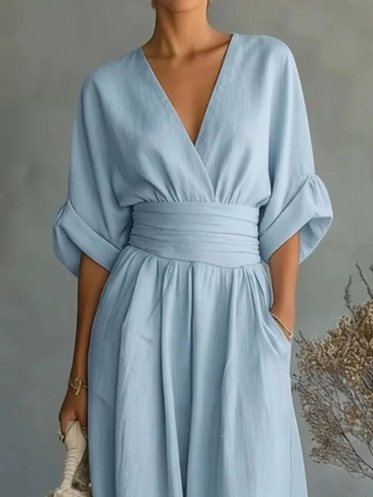 A woman wearing a light blue midi dress with a wrap-style V-neckline, elbow-length puff sleeves, a matching fabric waist tie, and side pockets, photographed against a soft gray wall with dried floral decor.