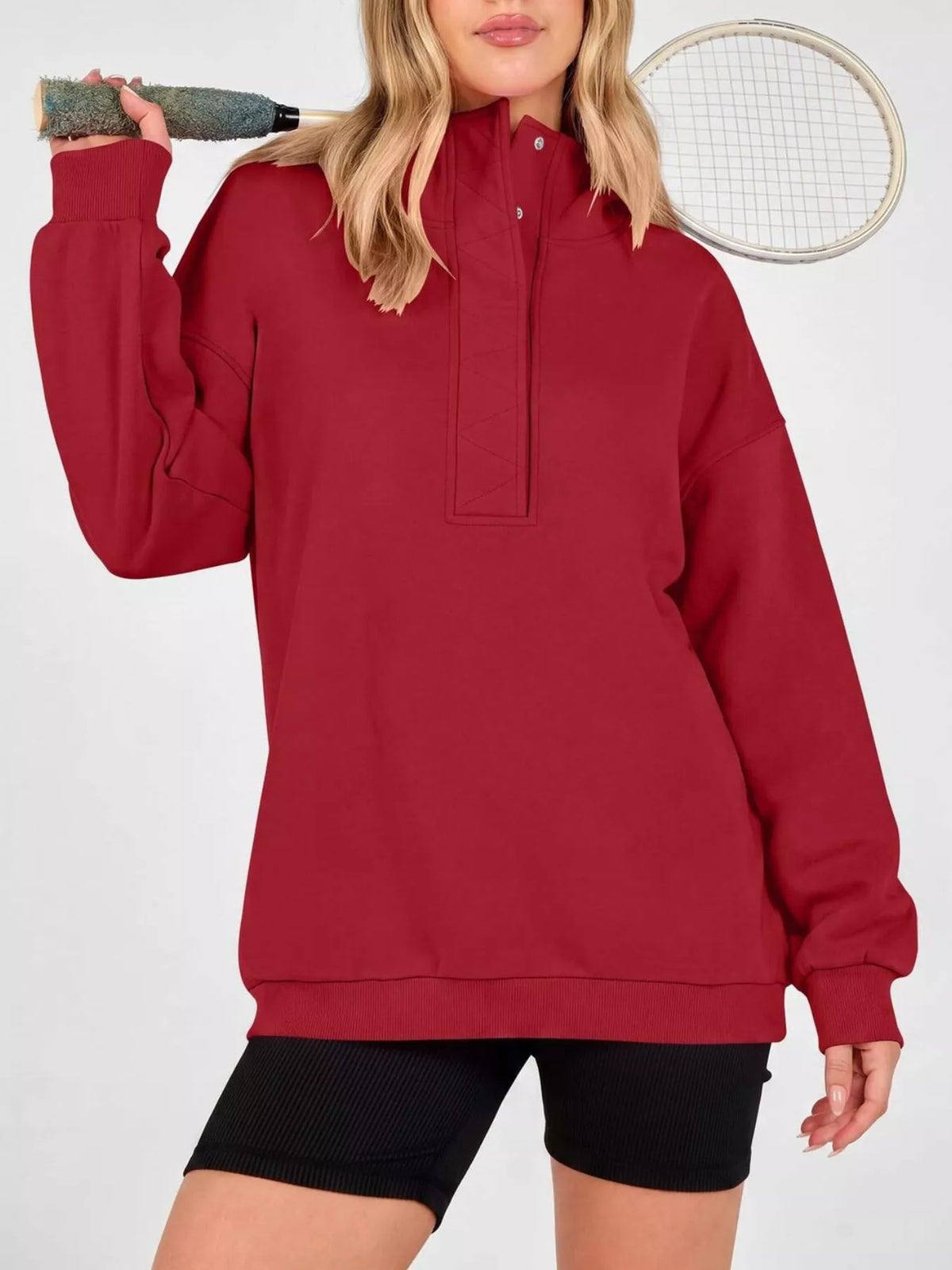 A woman wearing an oversized long-sleeve half-placket sweatshirt in deep red, paired with black athletic shorts, holding a tennis racket over her shoulder against a plain white background.