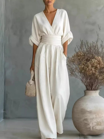 Woman in a white dress standing next to a vase with dried flowers against a gray wall.