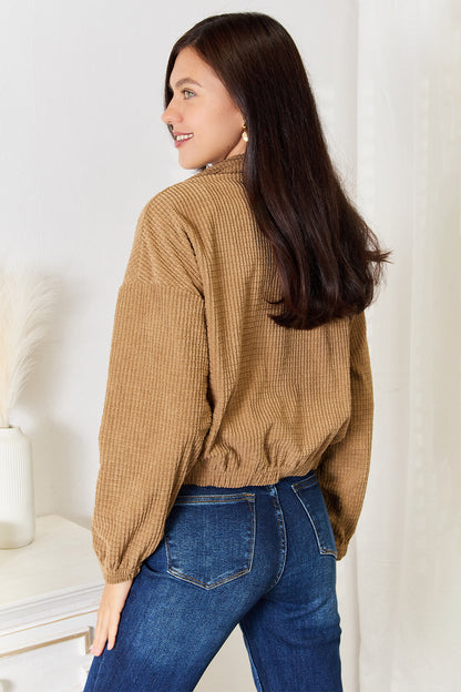Woman wearing a brown ribbed sweater and blue jeans against a white wall.