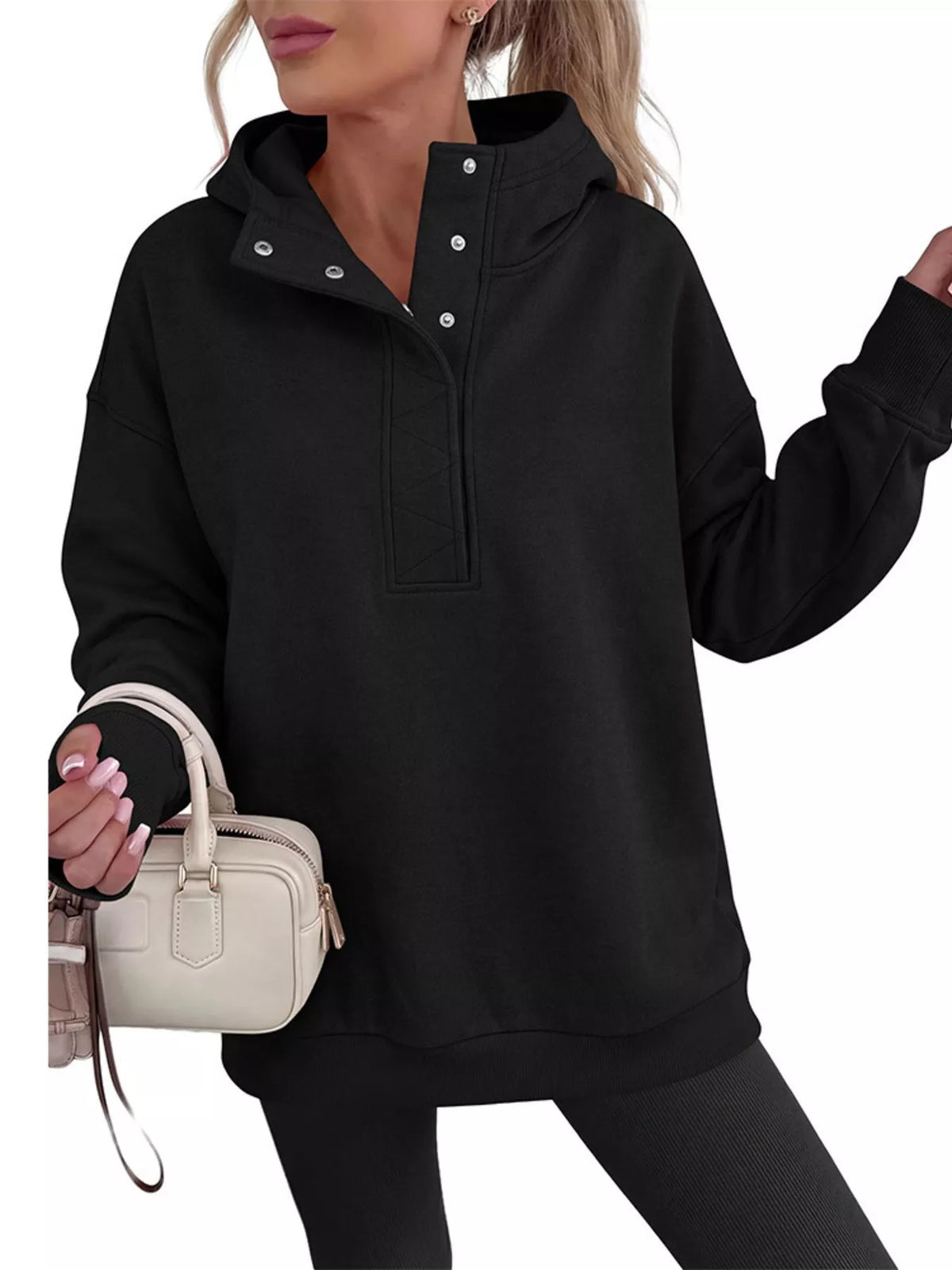 A woman wearing an oversized black pullover hoodie with a half-placket snap collar and drawstring hood, paired with black leggings and holding a small cream handbag against a plain white background.