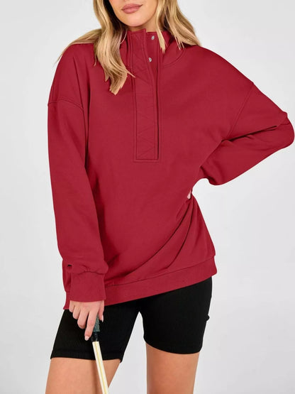 A woman wearing an oversized long-sleeve half-zip pullover in deep burgundy red, paired with black bike shorts, against a light gray studio background.