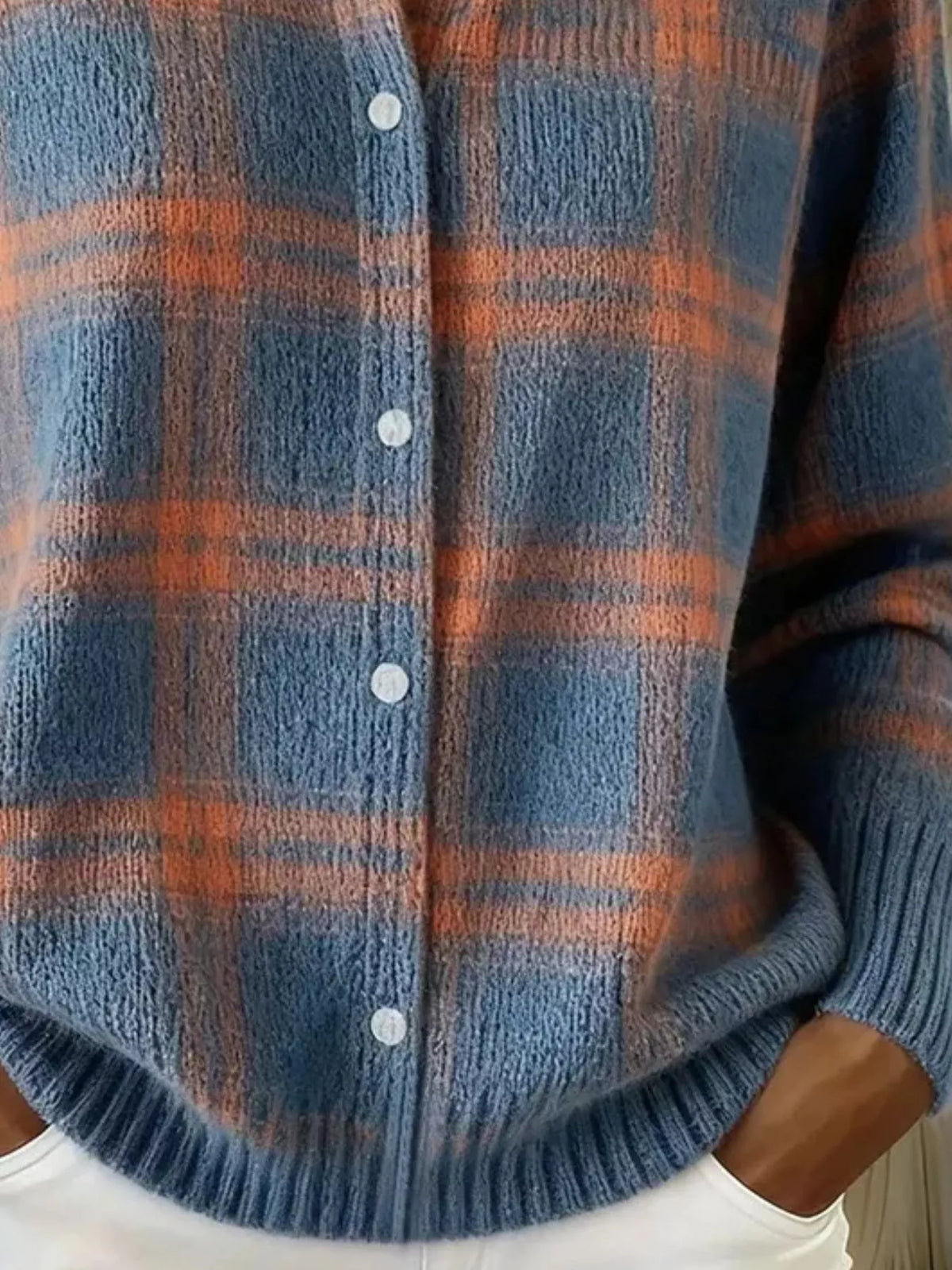 A close-up of a person wearing a fuzzy knit button-front cardigan with a blue and rust-orange plaid pattern, paired with white pants.