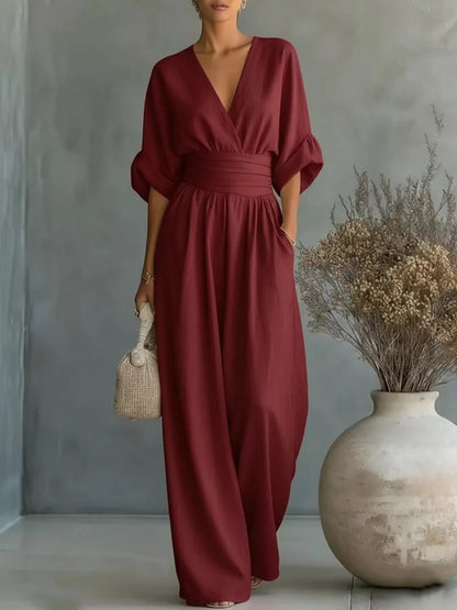 Woman wearing a burgundy jumpsuit holding a woven bag against a gray wall with a vase of dried flowers.