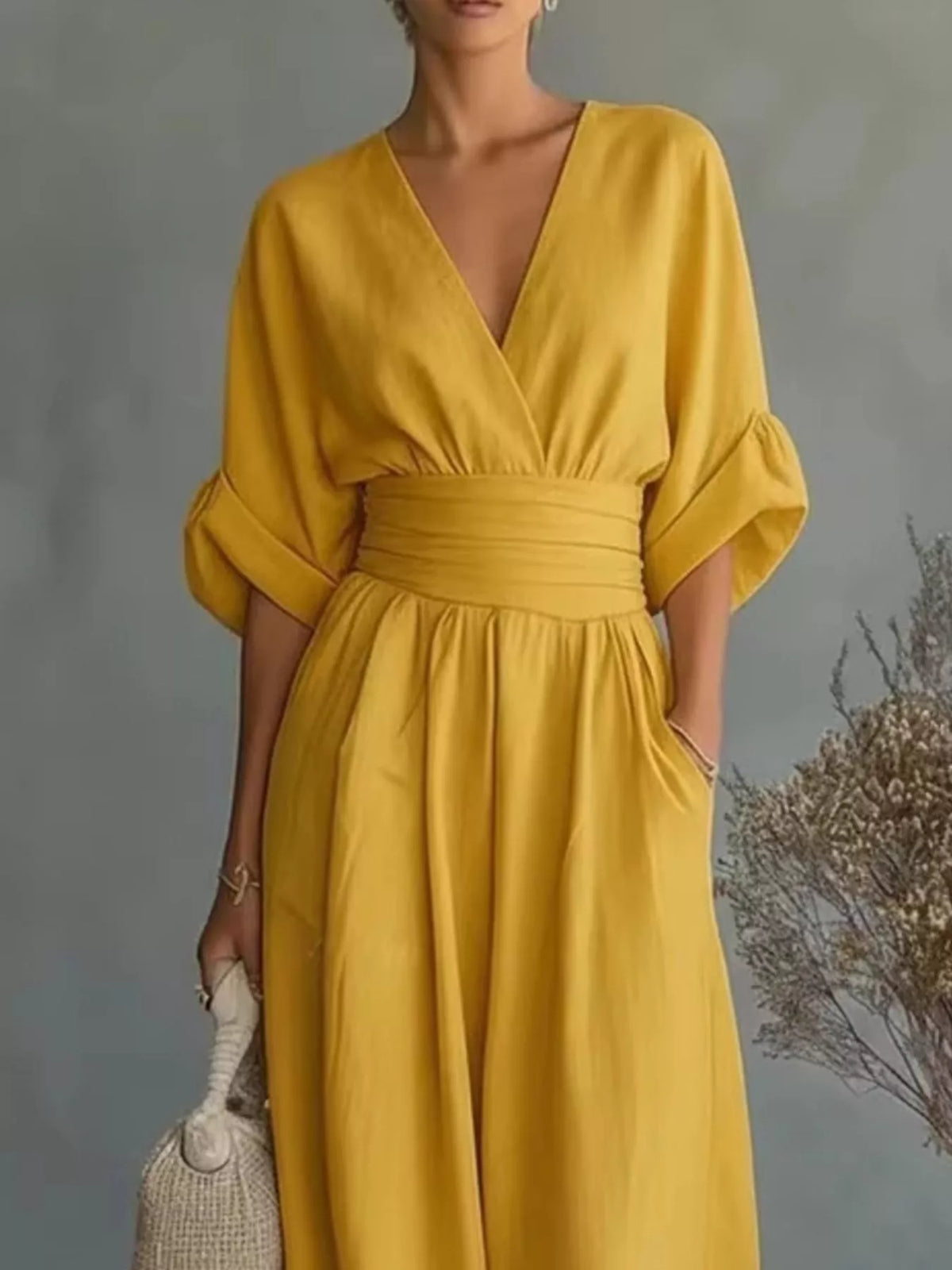 Woman wearing a yellow jumpsuit standing next to a vase with dried flowers against a gray wall.