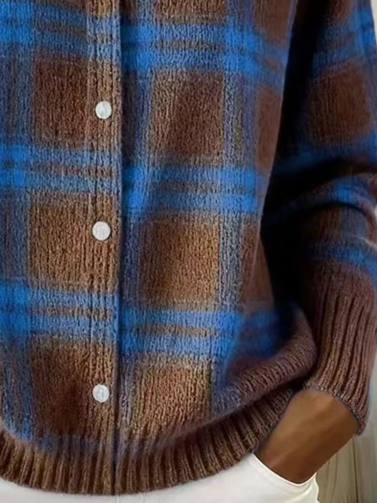 Close-up of a brown and blue plaid knit cardigan sweater with white buttons, worn by a person whose hand is visible in a white pocket.