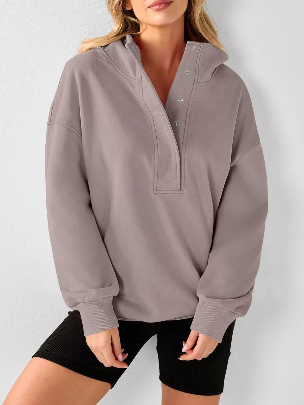A woman wearing an oversized taupe-gray pullover hoodie with a buttoned split neckline and ribbed cuffs, paired with black shorts, against a light gray studio background.