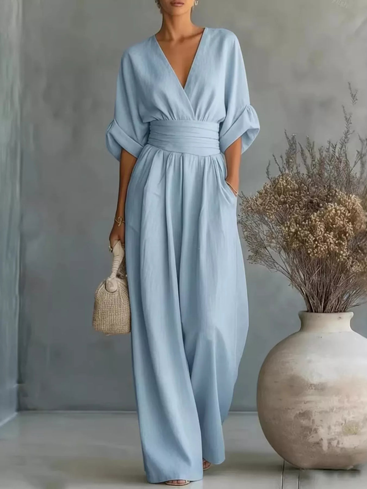 A woman models a light blue wrap-style jumpsuit with wide legs, elbow-length sleeves, and a matching fabric waist belt. She is standing in a studio setting with a textured gray wall, holding a small woven handbag, with a large ceramic vase of dried flowers on the right.
