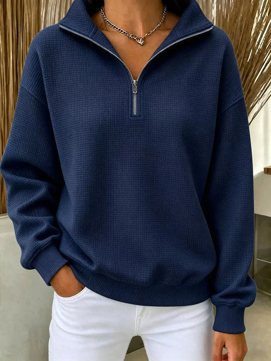 A person wearing a navy blue waffle-knit quarter-zip pullover sweatshirt with a stand collar and white pants, shown from the neck down in an indoor setting.