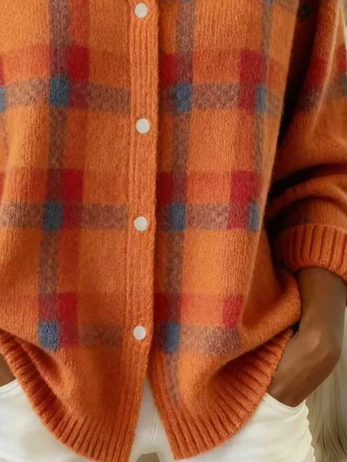 Close-up of a person wearing a fuzzy orange knit cardigan with a red and blue plaid pattern, white buttons, and ribbed cuffs and hem.