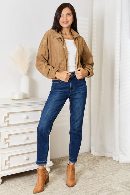 Woman wearing a brown jacket, white top, blue jeans, and brown boots in a room with a white dresser and curtains.