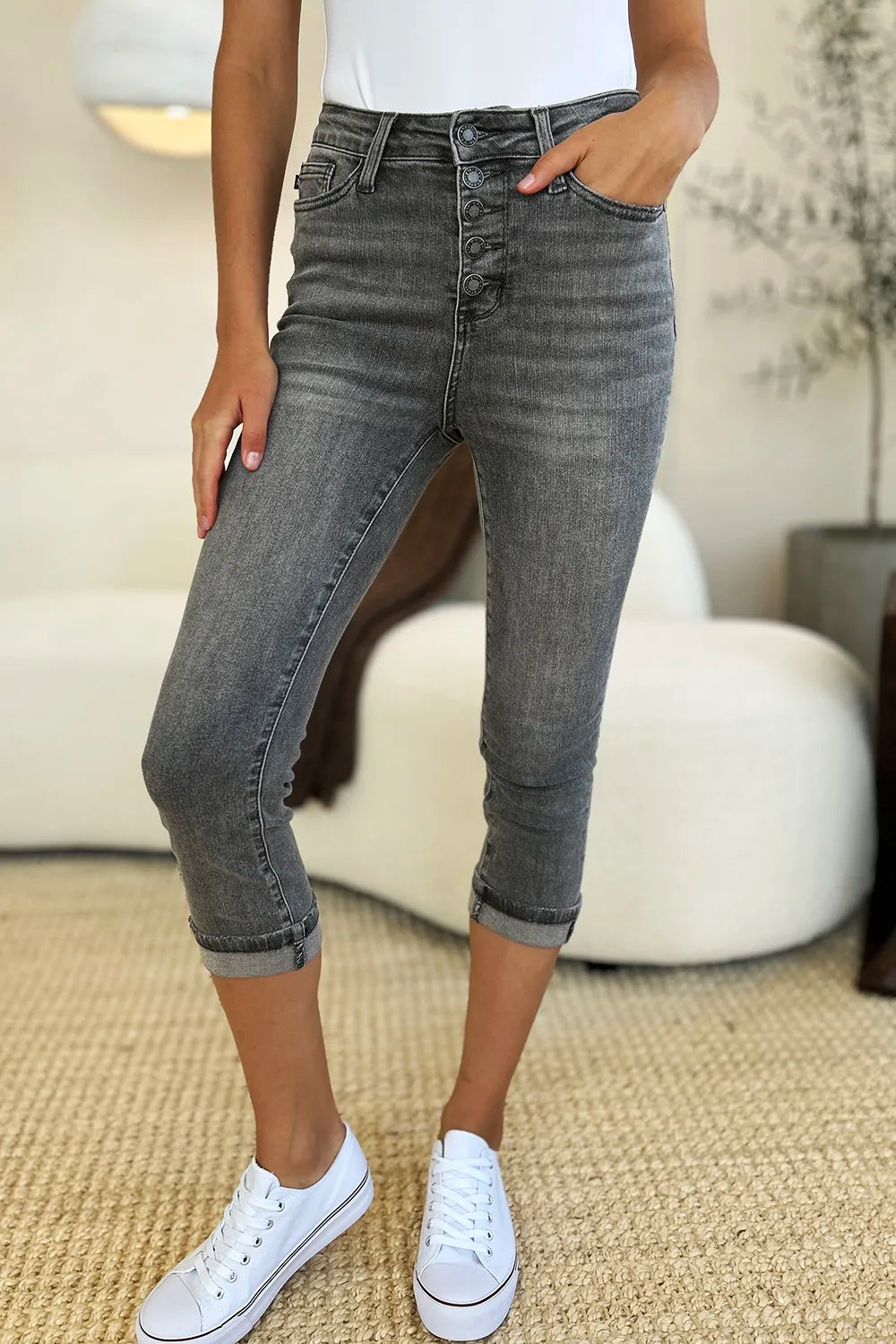 A person wearing dark gray high-waisted cropped jeans with a button fly, paired with a white top and white sneakers, standing in a bright living room.