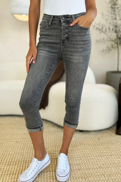 A person wearing dark gray high-waisted cropped jeans with a button fly, paired with a white top and white sneakers, standing in a bright living room.