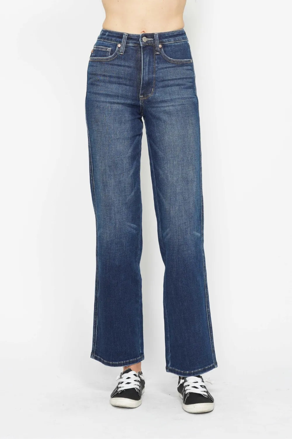 A person modeling a pair of high-waisted, wide-leg blue denim jeans with black and white sneakers, shown from the waist down against a plain white studio background.