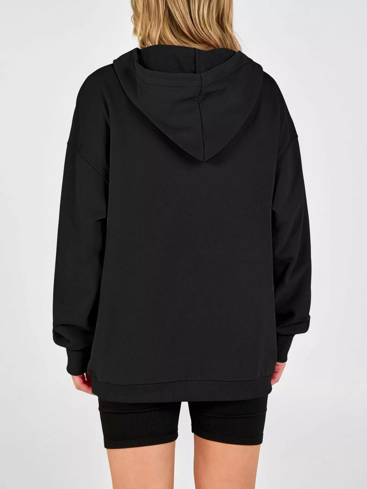 A person shown from the back wearing an oversized black pullover hoodie with a hood and long sleeves, paired with black fitted shorts, against a plain light gray studio background.