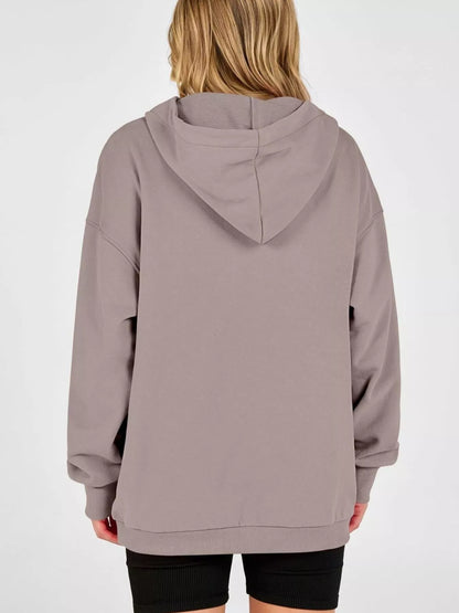 A person shown from the back wearing an oversized hooded sweatshirt in a muted taupe-gray color, paired with black shorts, against a plain light gray studio background.