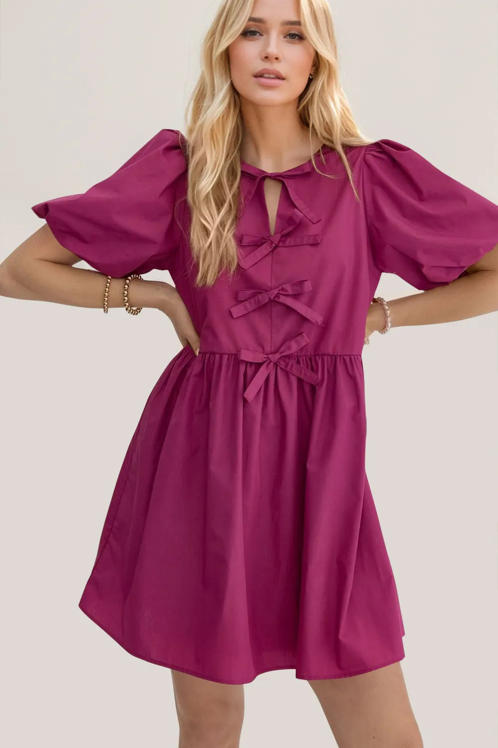 A woman modeling a short magenta puff-sleeve dress with a tie-neck detail and bow accents, photographed against a plain light beige studio background.