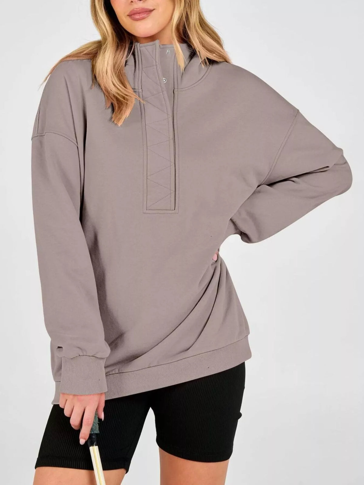 A woman wearing an oversized taupe gray mock-neck sweatshirt with a buttoned placket and black bike shorts, holding a golf club against a light gray studio background.