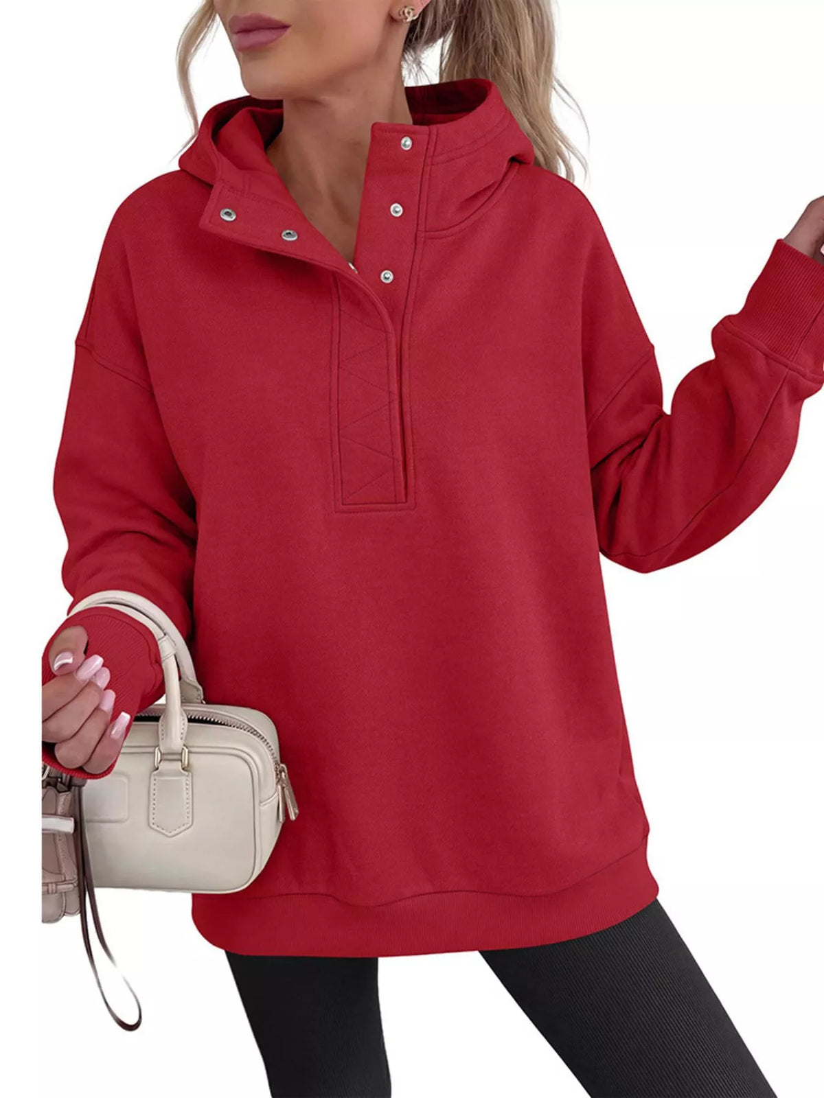 A woman wearing an oversized red pullover hoodie with a mock neck and snap-button placket, holding a small cream handbag against a plain white background.
