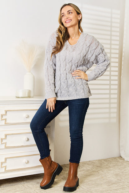 Woman wearing a gray cable knit sweater and blue jeans, standing in a room with a white dresser.