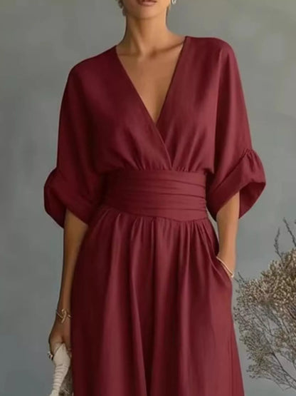 A woman wearing a deep burgundy wrap-style dress with a V-neckline, gathered waist, and elbow-length puff sleeves, standing against a soft gray studio background.