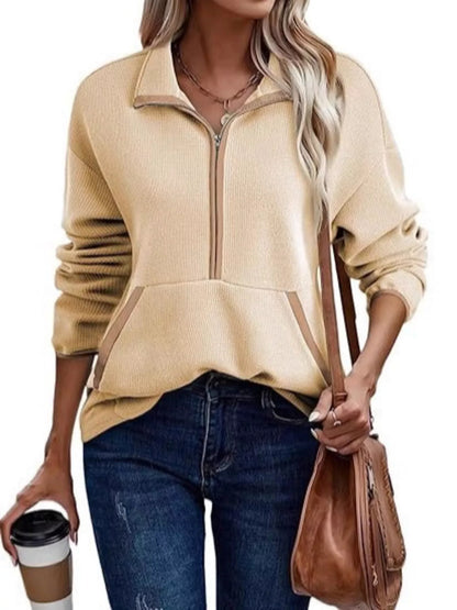 Woman wearing a beige zip-up sweater with blue jeans, holding a brown bag and a coffee cup.