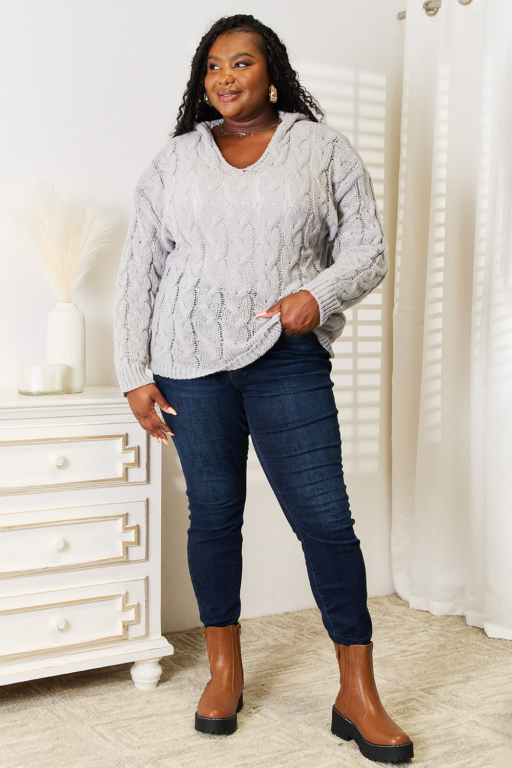 Woman wearing a gray cable knit sweater and blue jeans standing in a room with white furniture.