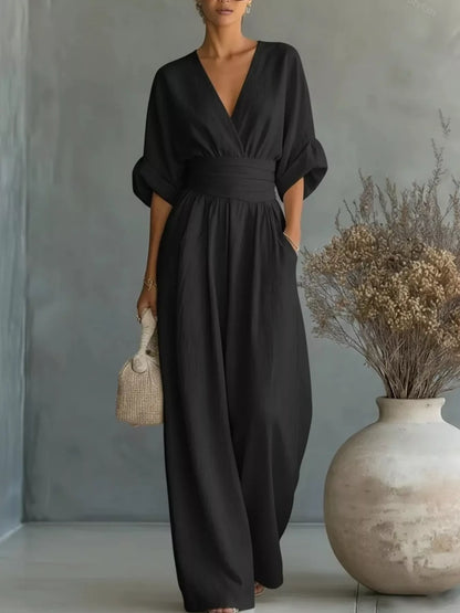 Woman wearing a black jumpsuit standing next to a vase with dried plants against a gray wall.