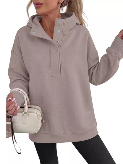 A woman wearing an oversized taupe pullover hoodie with a mock neck and snap details, holding a small cream handbag against a plain white background.