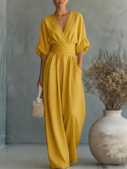 Woman wearing a yellow jumpsuit standing next to a vase with dried flowers against a gray wall.