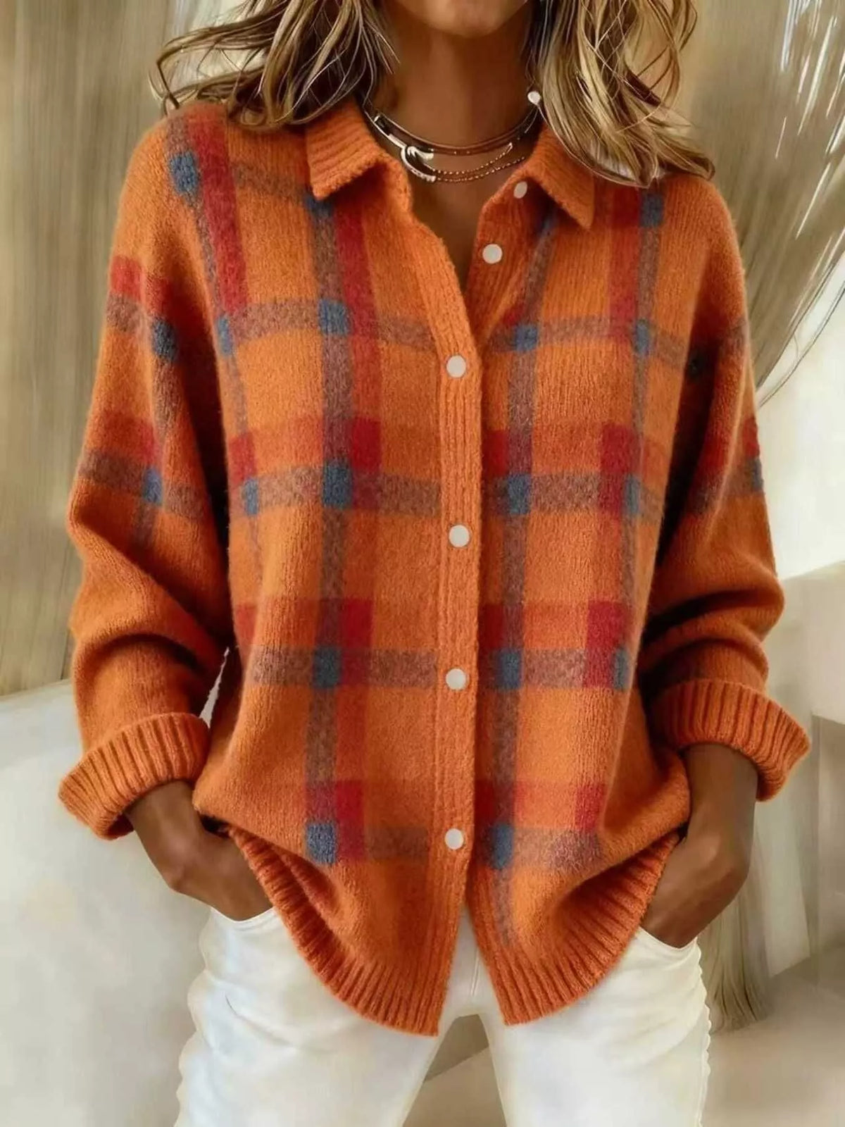 A woman wearing a fuzzy orange knit button-front cardigan with a red and blue plaid pattern, paired with white pants and layered gold necklaces.