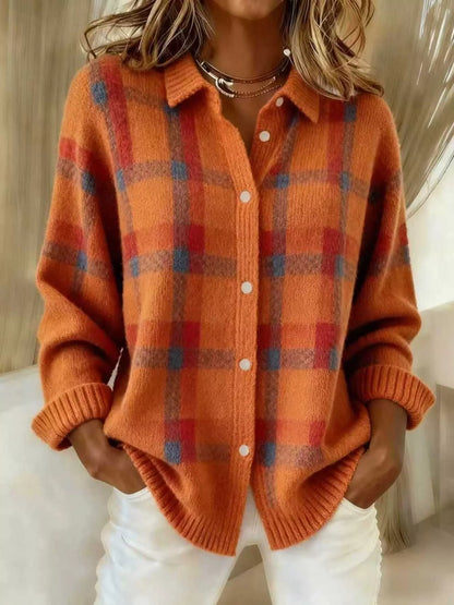 A woman wearing a fuzzy orange knit button-front cardigan with a red and blue plaid pattern, paired with white pants and layered gold necklaces.